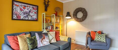 A living room with a grey sofa, a blue armchair, a wooden side table with oranges, a bookshelf, a wall-mounted wreath, and a chandelier.