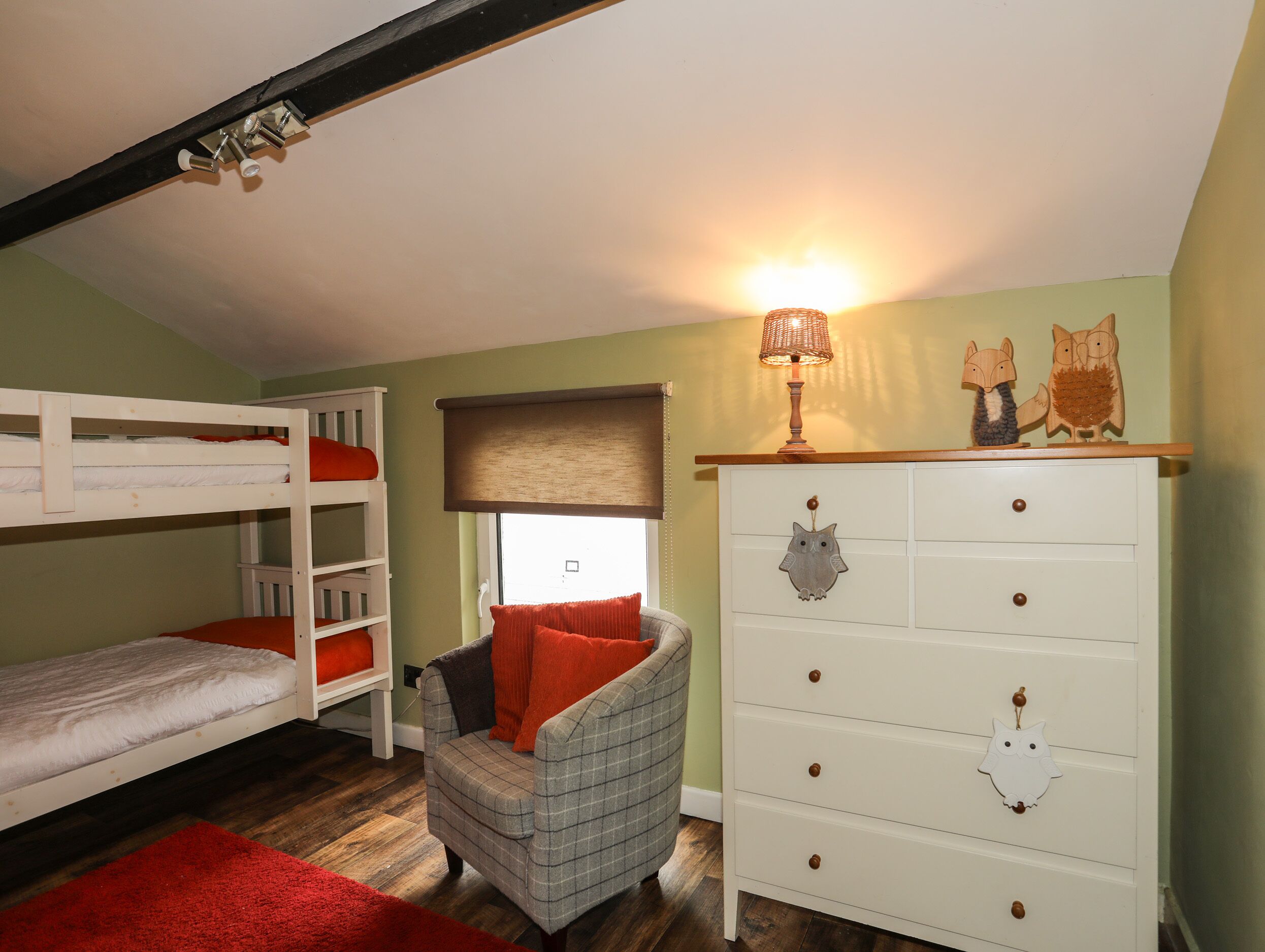 Ein Etagenbett mit einem roten Kopfkissen, ein Karo-Sessel, eine weiße Kommode mit Eulen-Dekorationen und eine Lampe darauf.