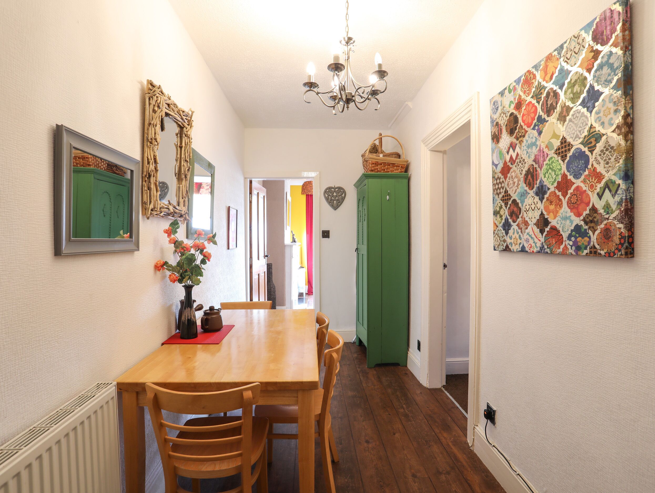 Un coin repas avec une table en bois, des chaises, un vase de fleurs, un meuble vert et une décoration murale colorée.