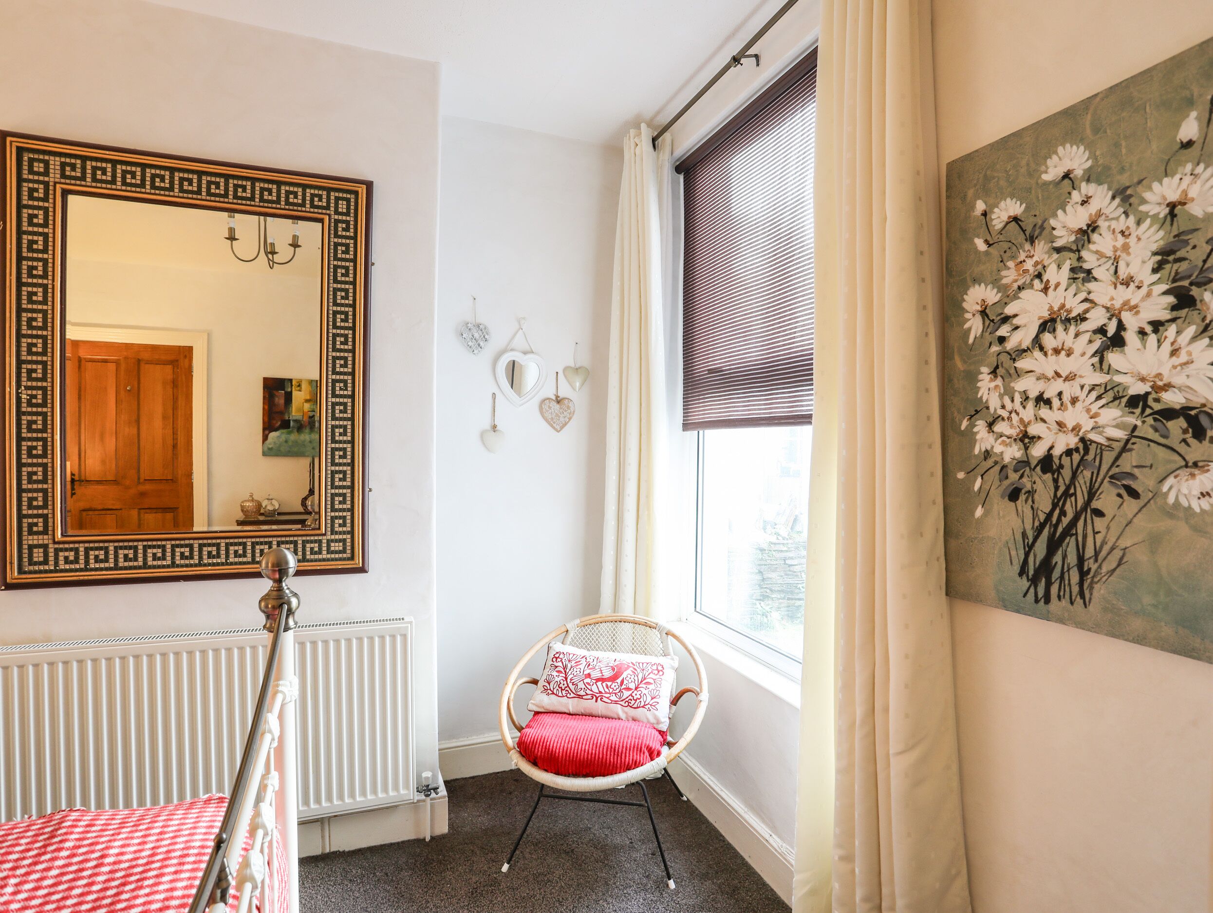 Une pièce avec un grand miroir, un radiateur, un fauteuil en osier avec un coussin rouge et une fenêtre avec des stores.