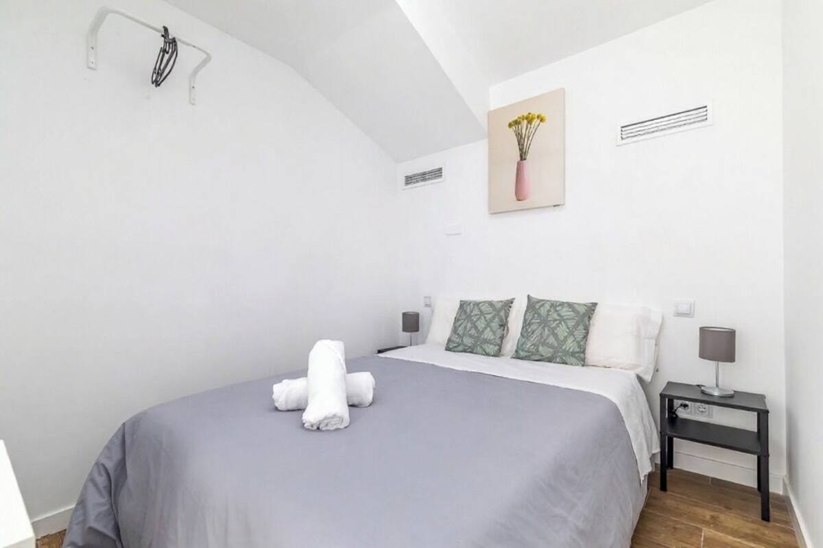 A modern bedroom with a large bed, a bedside table with a lamp, and a wall-mounted air vent.