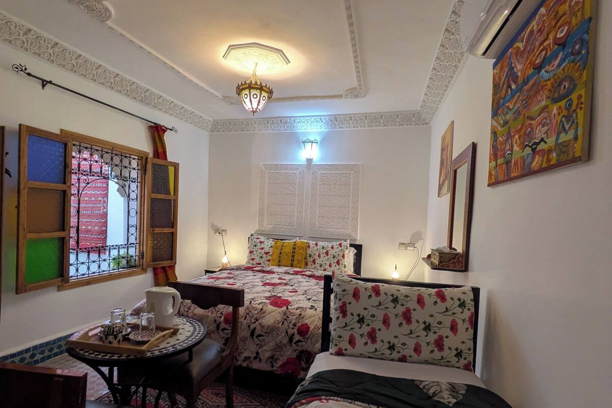 A room with a bed, a table with a chair, a window with decorative shutters, and a ceiling light fixture.