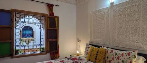 Un dormitorio con cubrecama floral, una lámpara decorativa en el techo, una ventana con intrincados detalles de hierro y adornos murales estampados.