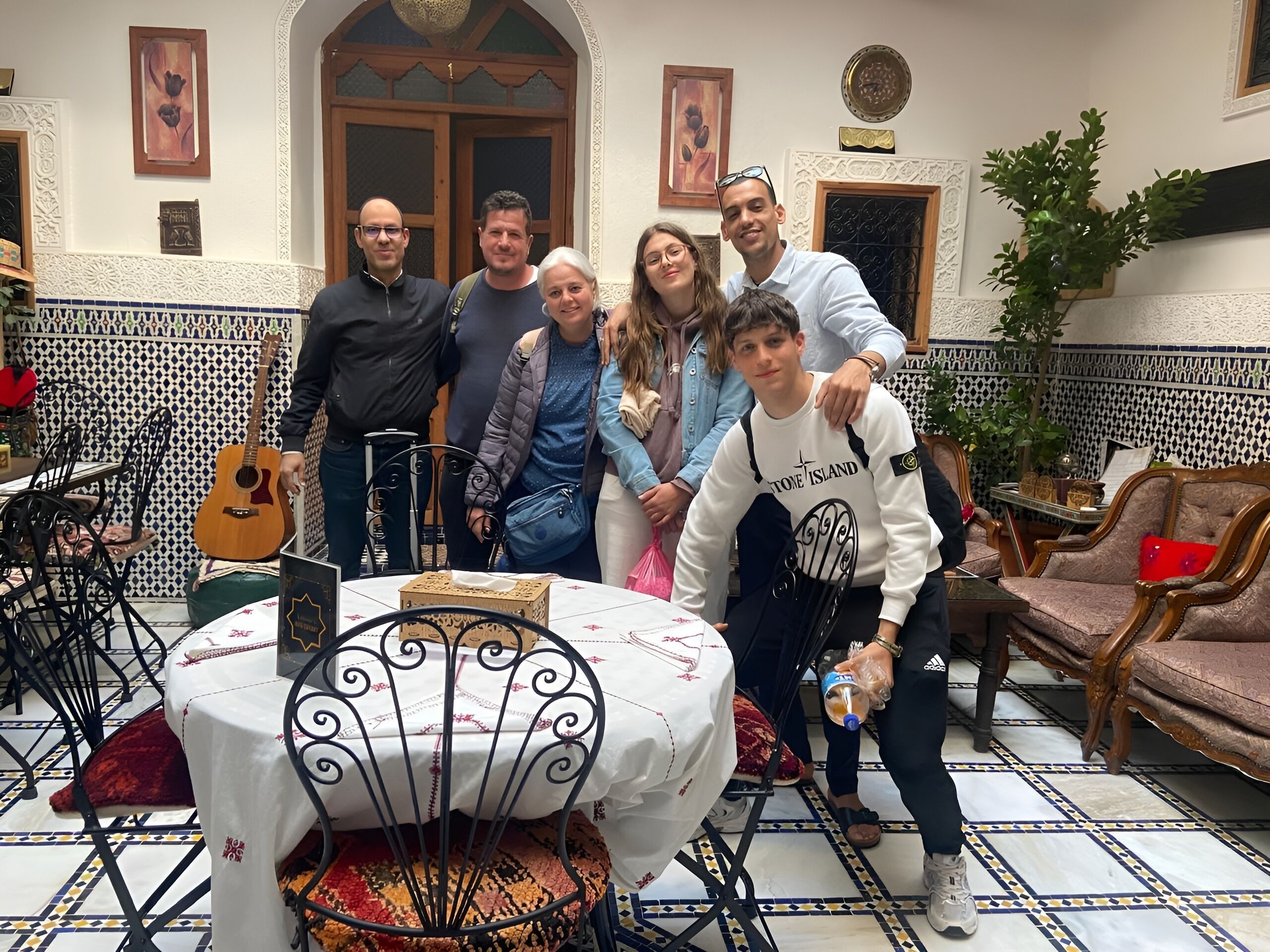 A group of people gathered around a table with a white tablecloth, in a room with patterned tile flooring and traditional Moroccan-style decor.