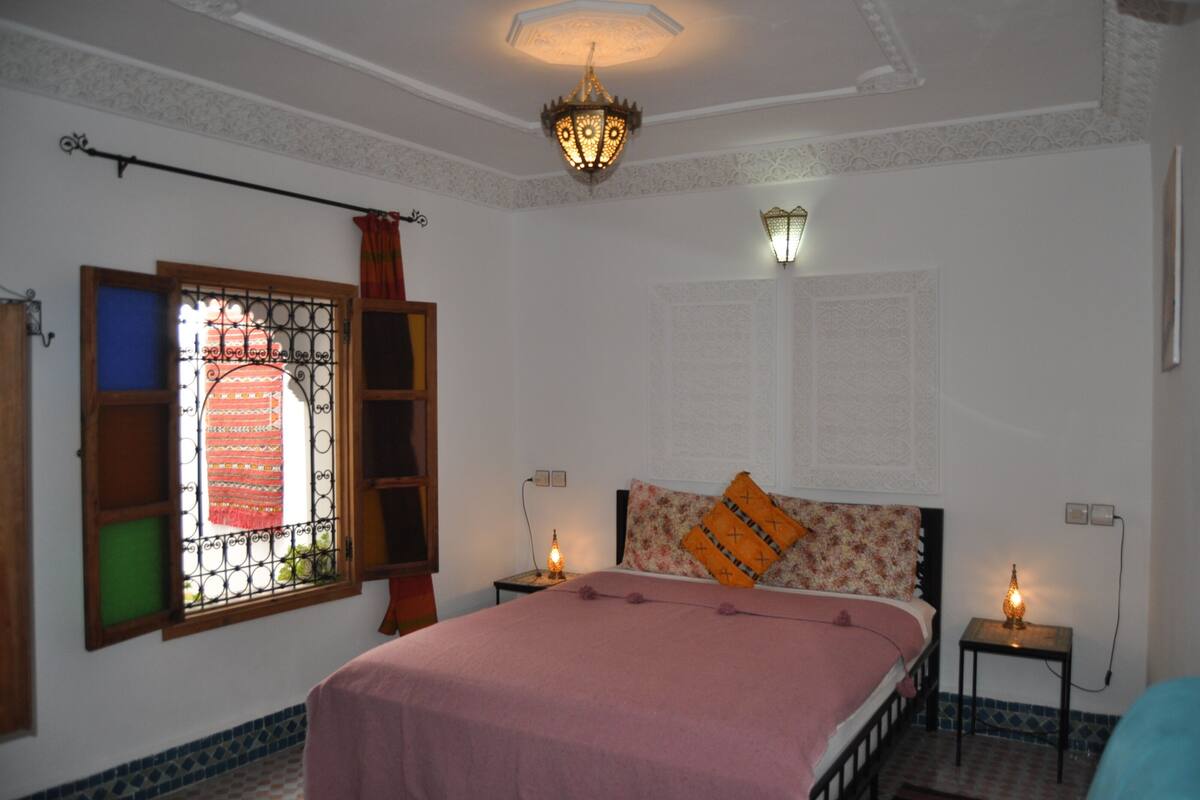 A bedroom with a bed, two bedside tables, a window with a decorative iron grill, and a chandelier.