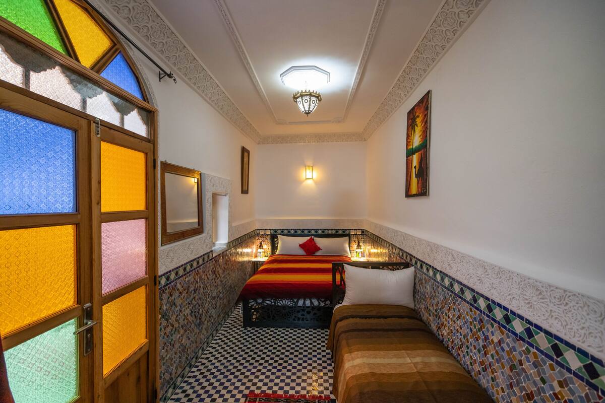 A room with a checkered floor, a bed with a red and black striped bedspread, and a wall-mounted light fixture.