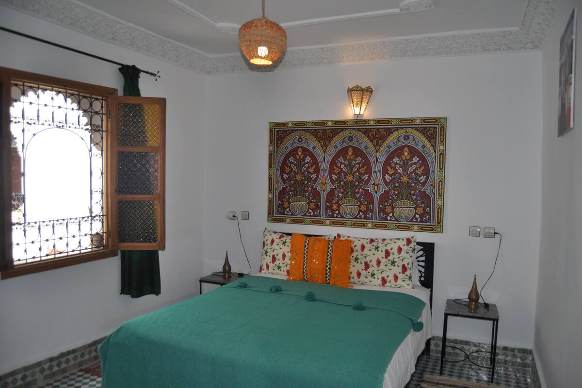 A bedroom with a bed, a decorative wall hanging, a window with a metal grill, and a hanging light fixture.