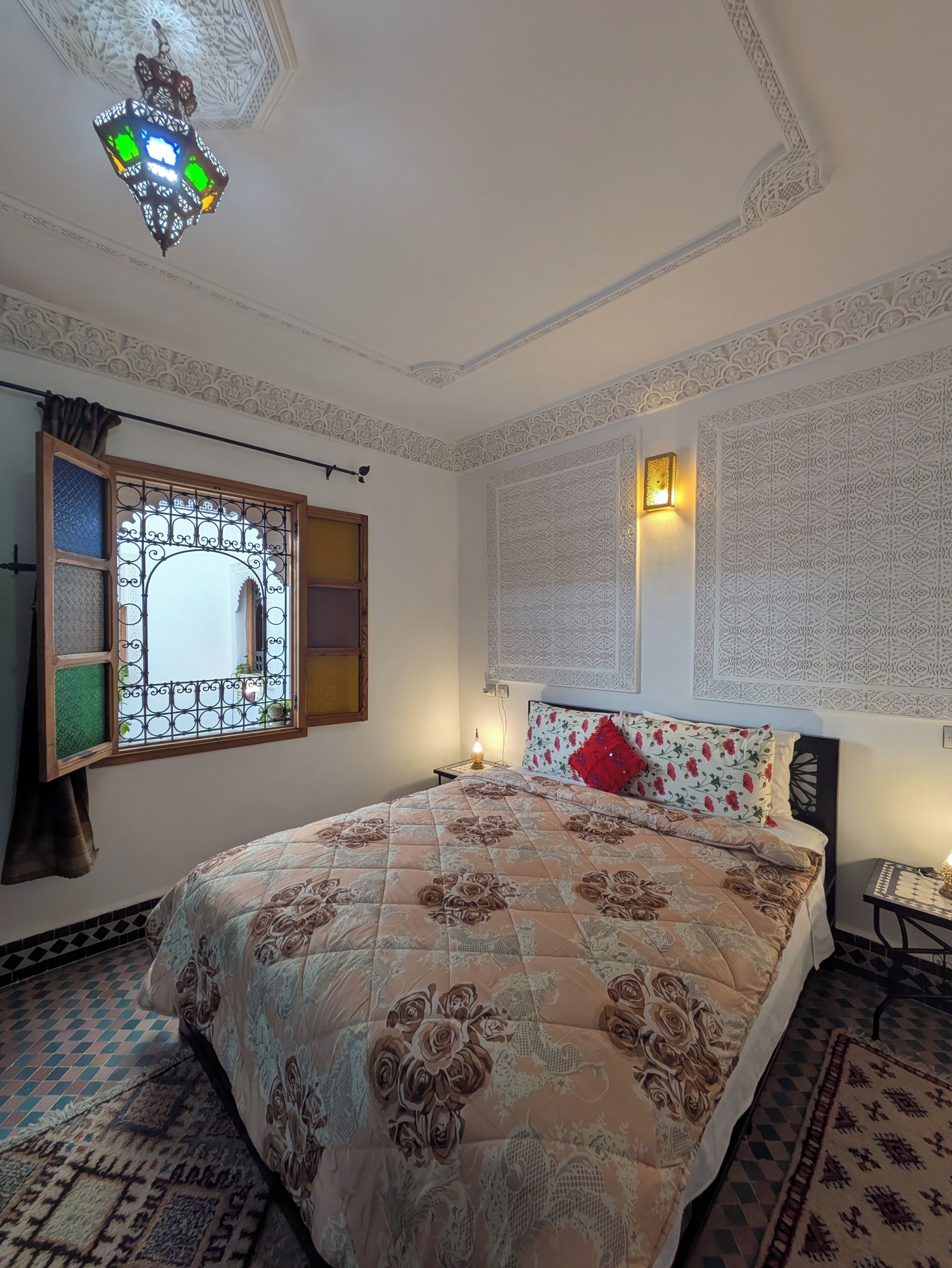 Un dormitorio con una colcha estampada, una lámpara de techo decorativa, una ventana con reja de metal y decoración mural con motivos.