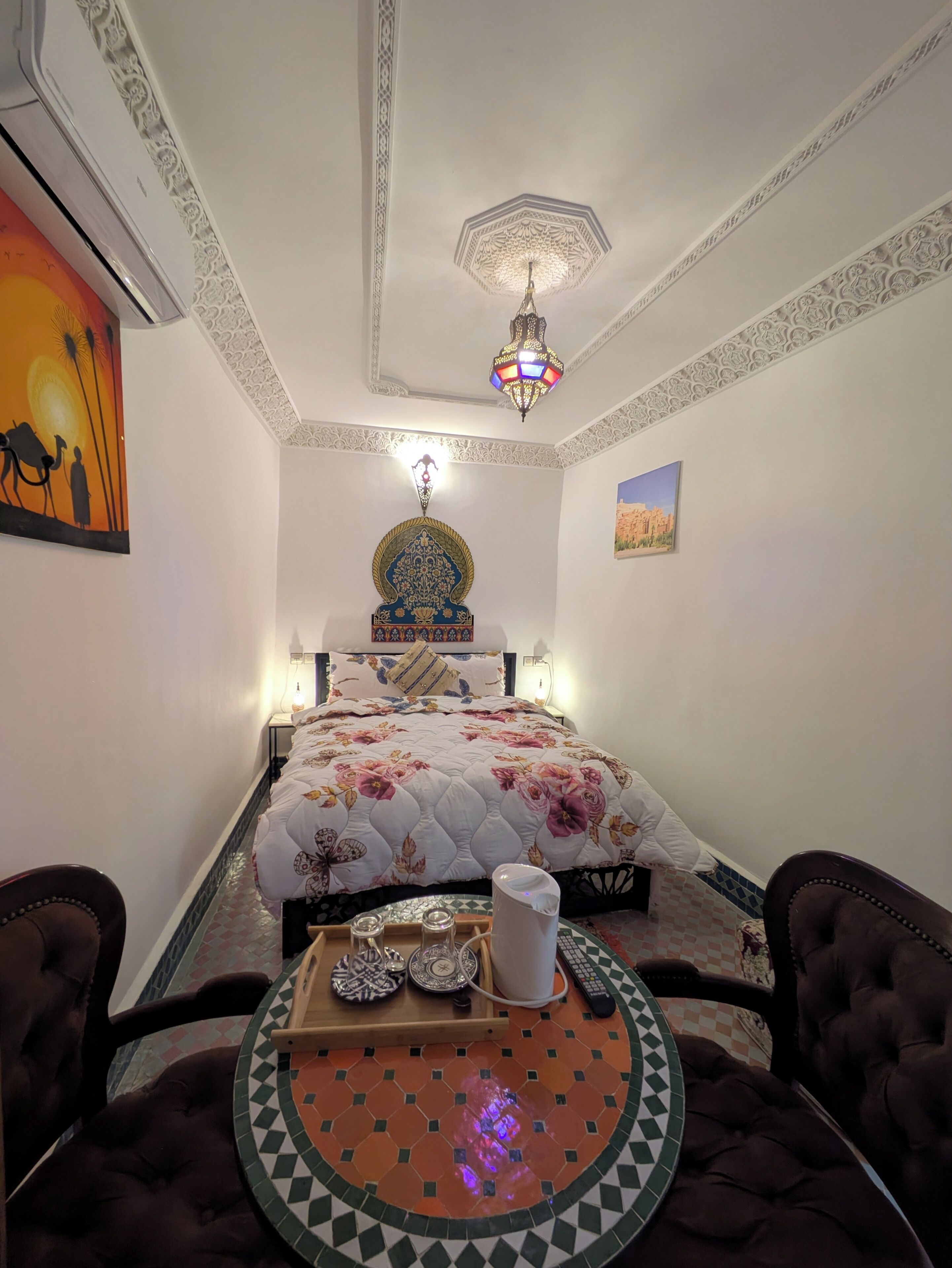 A room with a bed, a decorative ceiling light, a wall-mounted lamp, a wall painting, and a small table with a tray.