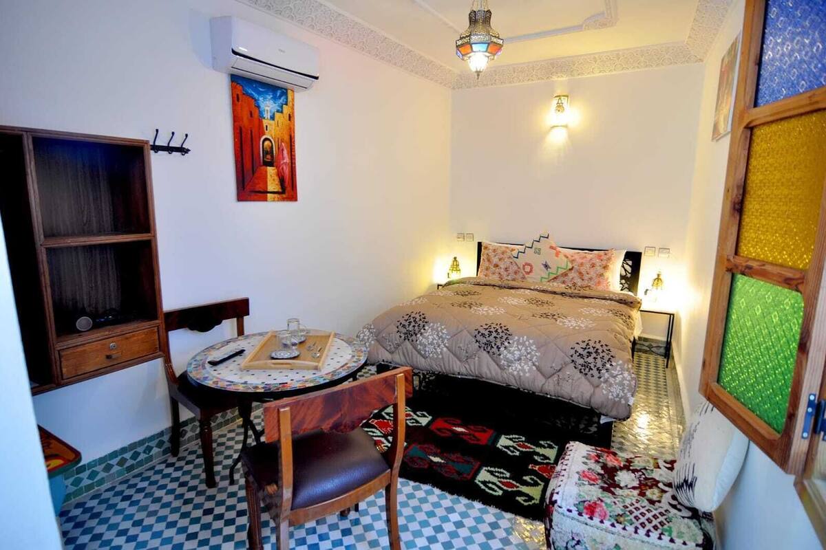 A bedroom with a bed, a small table with a tray, a chair, a wall-mounted air conditioner, and a decorative painting.
