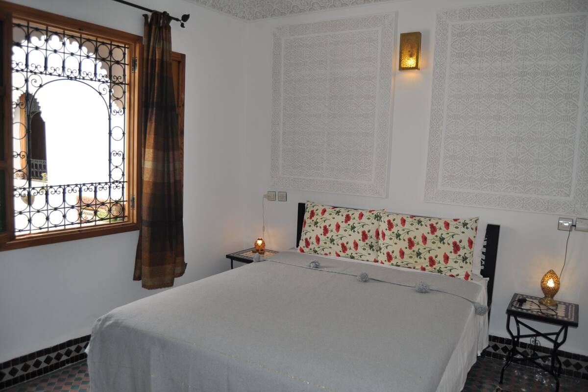 A bedroom with a bed, a window with curtains, a nightstand with a lamp, and decorative wall panels.