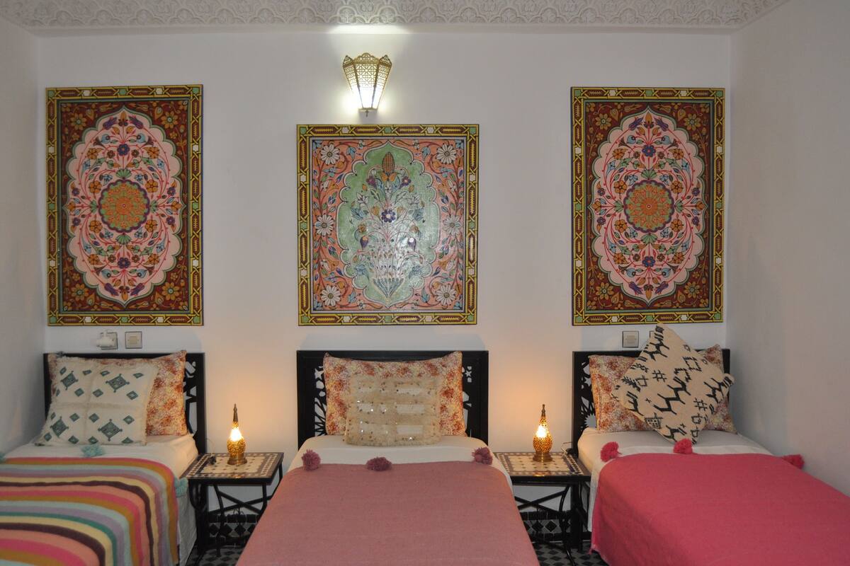 A room with two beds, each with a unique patterned headboard and decorative pillows. There are wall-mounted lamps and a large ornate tapestry above the beds.
