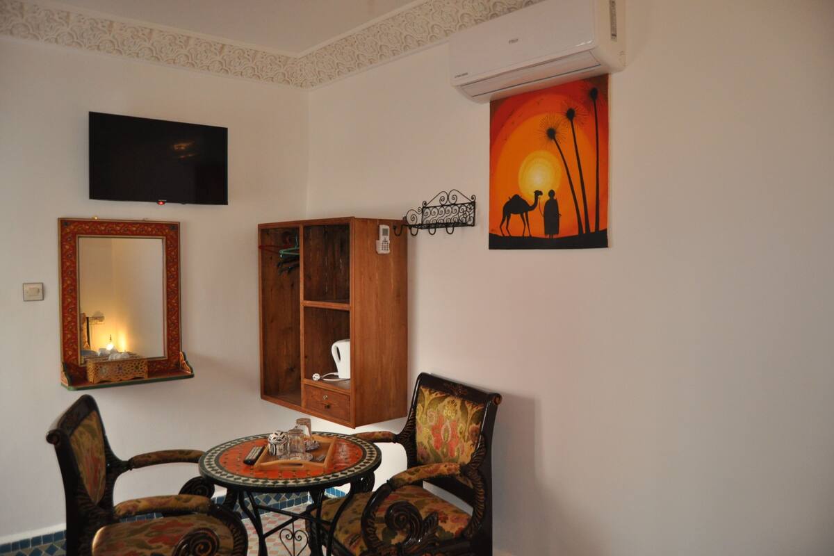A living room with a TV, a large mirror, a wooden cabinet, a round table with chairs, and a sunset painting.
