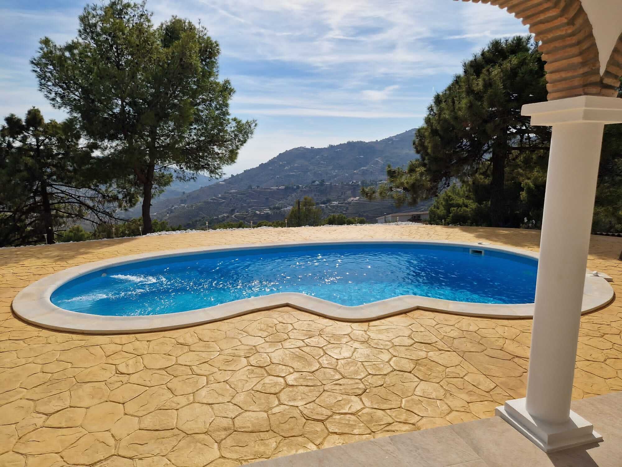 A swimming pool with a hexagonal tiled deck, surrounded by trees and a mountainous backdrop.