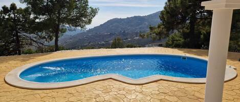 A swimming pool with a hexagonal tiled deck, surrounded by trees and a mountainous backdrop.