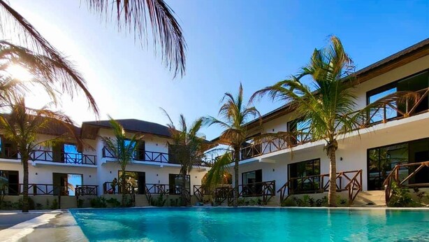 Una zona de piscina con agua cristalina, rodeada de palmeras y un edificio con balcones.