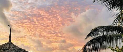 A beach with a thatched umbrella, two wooden lounge chairs, and a view of the ocean at sunset.