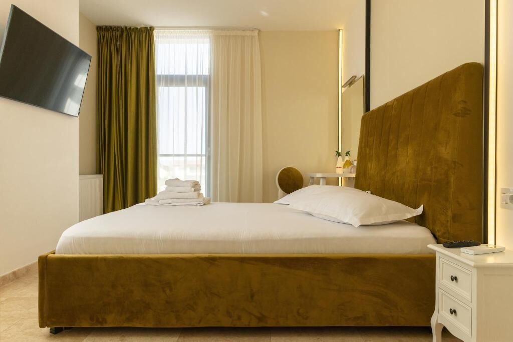 Habitación de hotel con una cama grande, una mesita de noche con lámpara, un televisor montado en la pared y una ventana con cortinas.