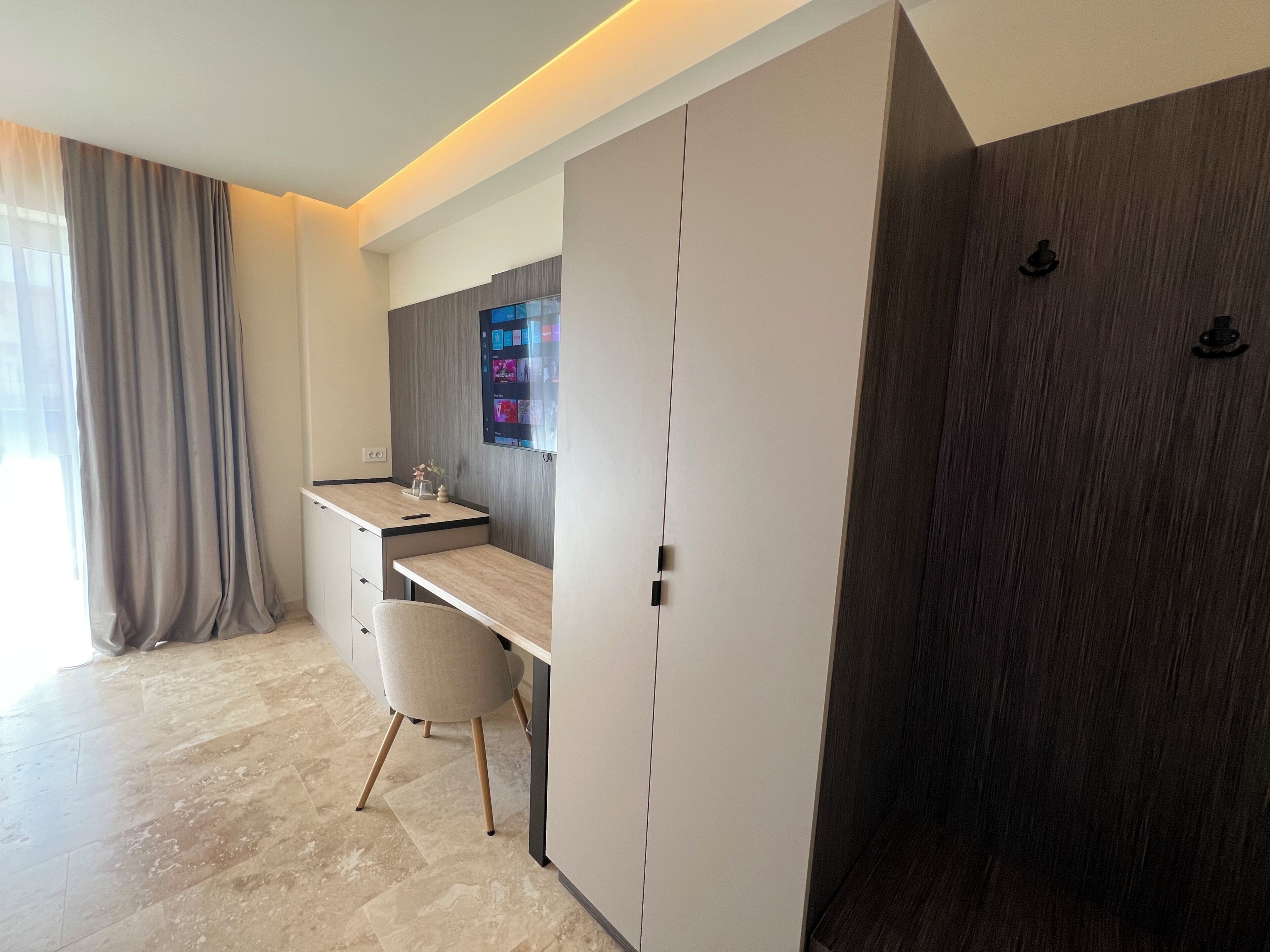 A modern bedroom with a built-in desk, a chair, a wardrobe, and a large window with curtains.