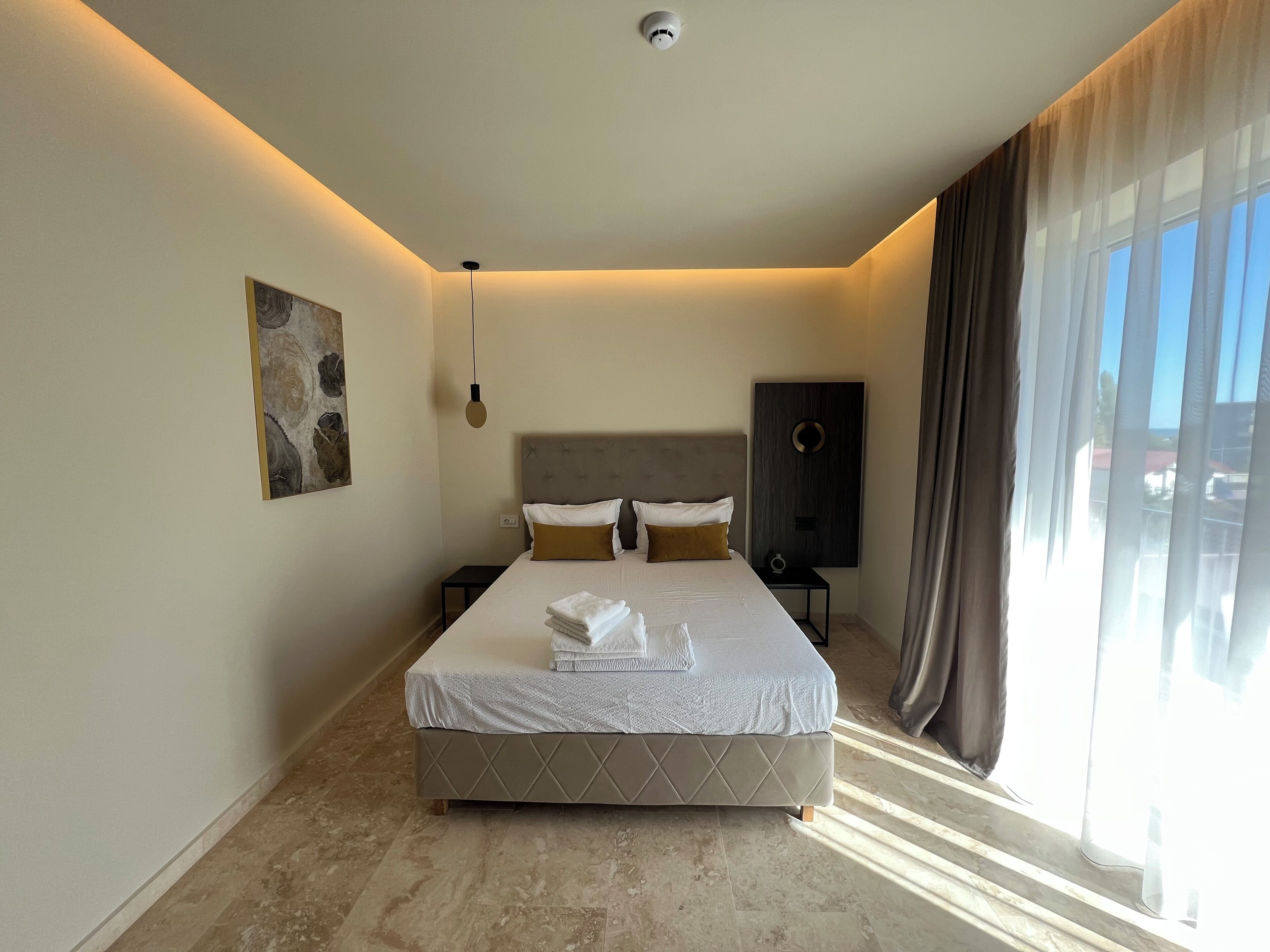 A modern hotel room with a large bed, a headboard, a bedside table, a wall-mounted artwork, and a view of the outside through sheer curtains.