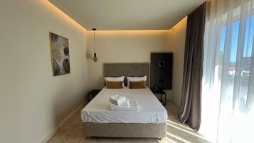 A modern hotel room with a large bed, a headboard, a bedside table, a wall-mounted artwork, and a view of the outside through sheer curtains.