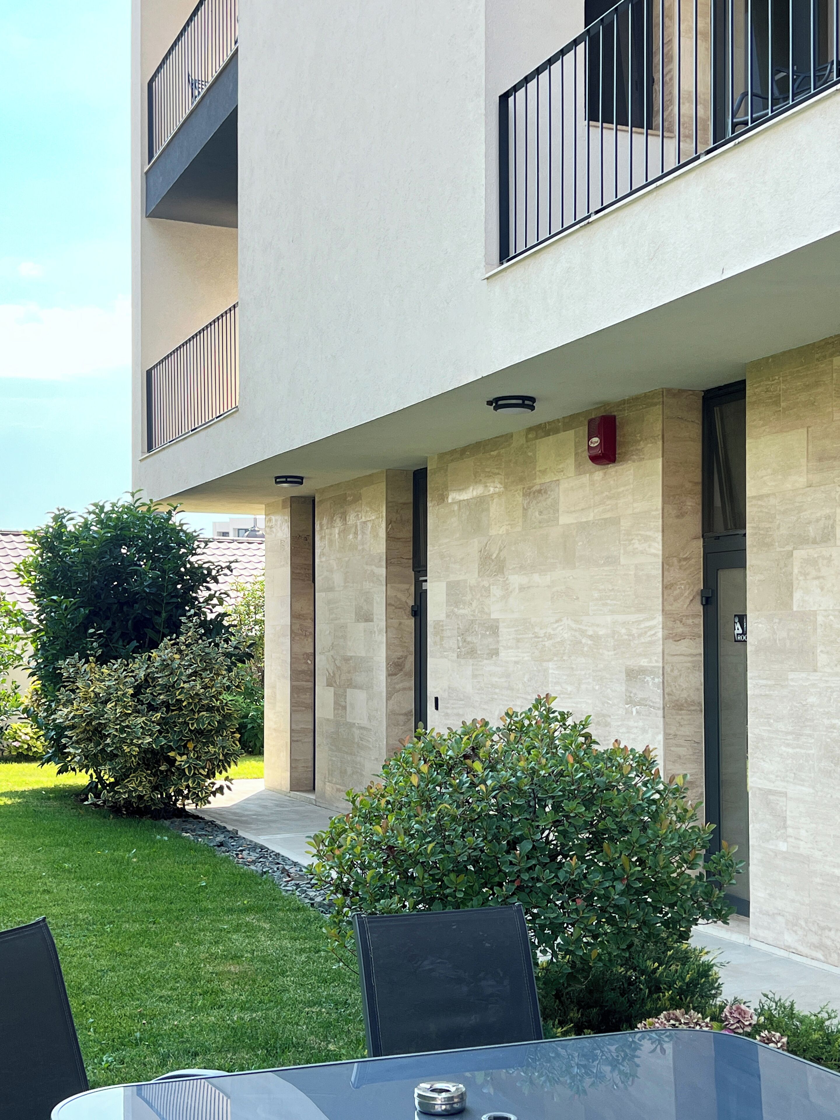 Um edifício moderno com telhado plano, uma área externa coberta com mesa de vidro e um pequeno jardim.