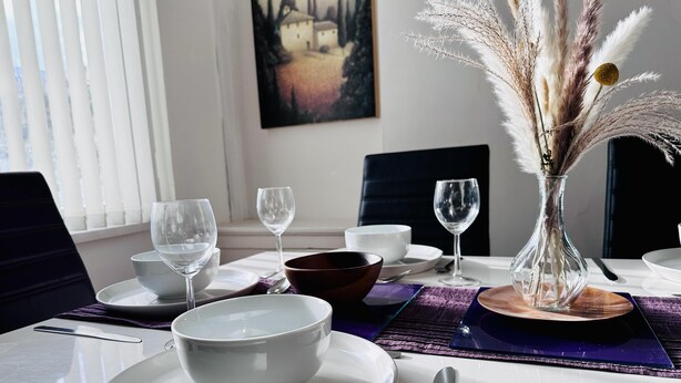Une table à manger dressée avec de l’assiette blanche, des bols et des verres à vin, un ordinateur portable et un vase contenant des plumes d’herbe de la pampa.