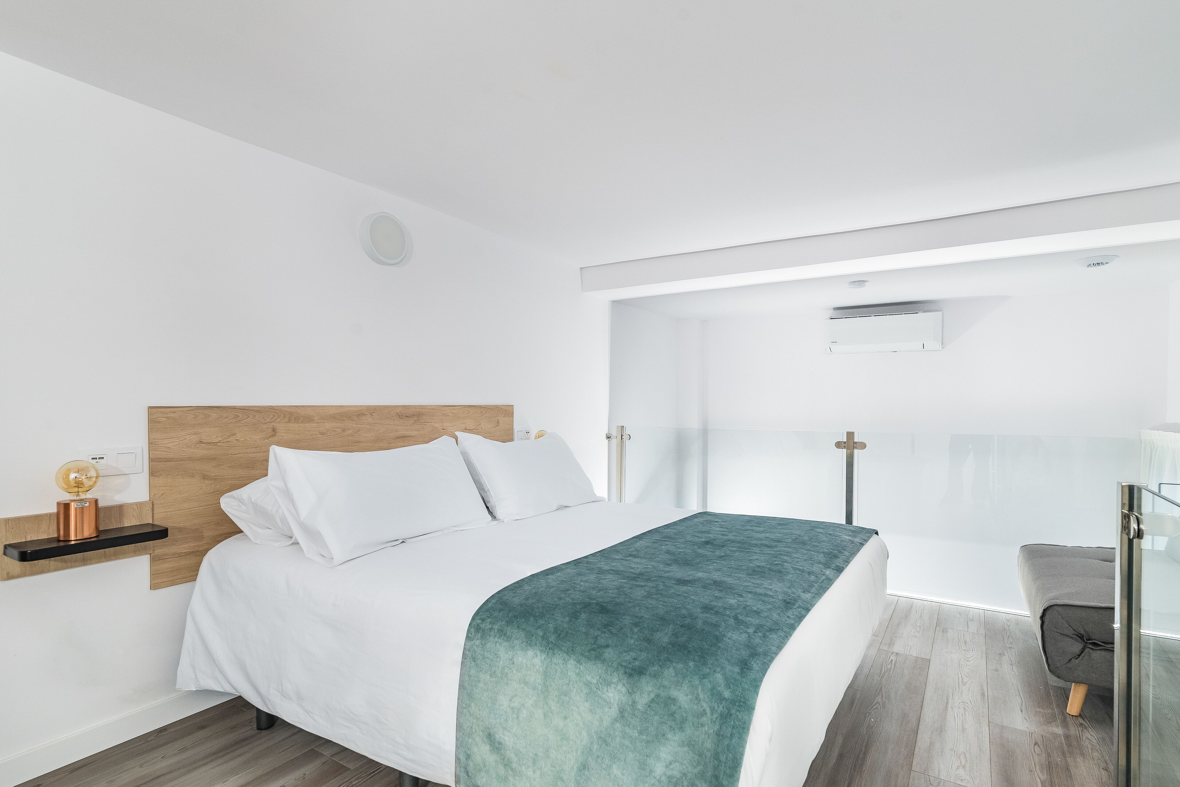 A modern bedroom with a wooden headboard, white bedding, a teal throw blanket, a bedside table with a lamp, and a glass door leading to a balcony.
