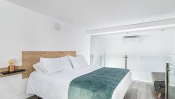 Un dormitorio moderno con cabecera de madera, ropa de cama blanca, una manta color turquesa, una mesita de noche con lámpara y una puerta de vidrio que da a un balcón.