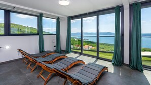 Una habitación con un gran ventanal corredero que da a un balcón con vistas a un lago y colinas. Hay cuatro sillones de madera con cojines grises.