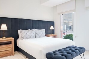Un dormitorio moderno con una cama grande, dos lámparas de noche, un banco y vistas de la ciudad a través de amplios ventanales.