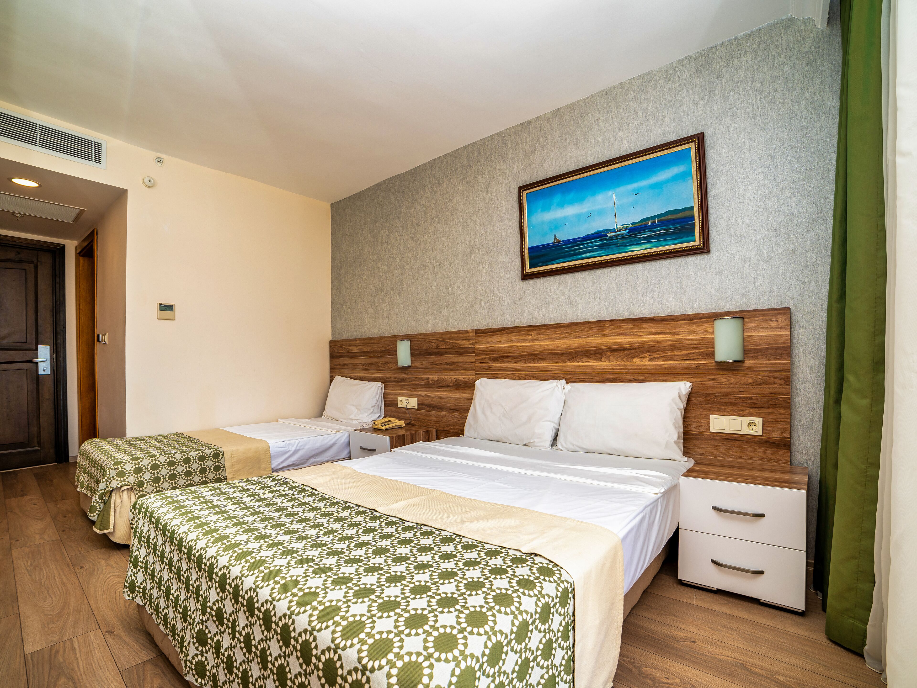 Habitación de hotel con dos camas, cabecero de madera, pintura de un paisaje marino y cortina verde.