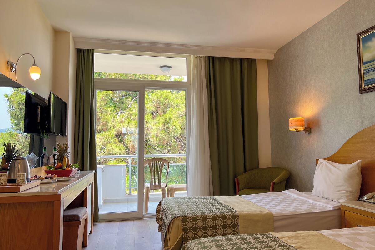 Habitación de hotel con dos camas, un escritorio con televisor, una silla y un balcón con cortinas.