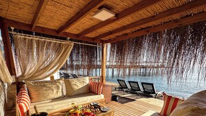 Un área de estar exterior lujosa con terraza de madera, asientos acolchados, una mesa con frutas y una botella, y vistas al agua.