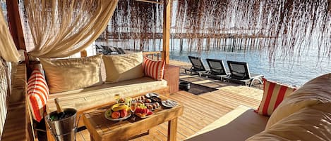 A luxurious outdoor seating area with a wooden deck, cushioned seating, a table with fruits and a bottle, and a view of the water.