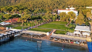 Vista aérea de un complejo turístico costero con un puerto deportivo, asientos en la playa y montañas al fondo.