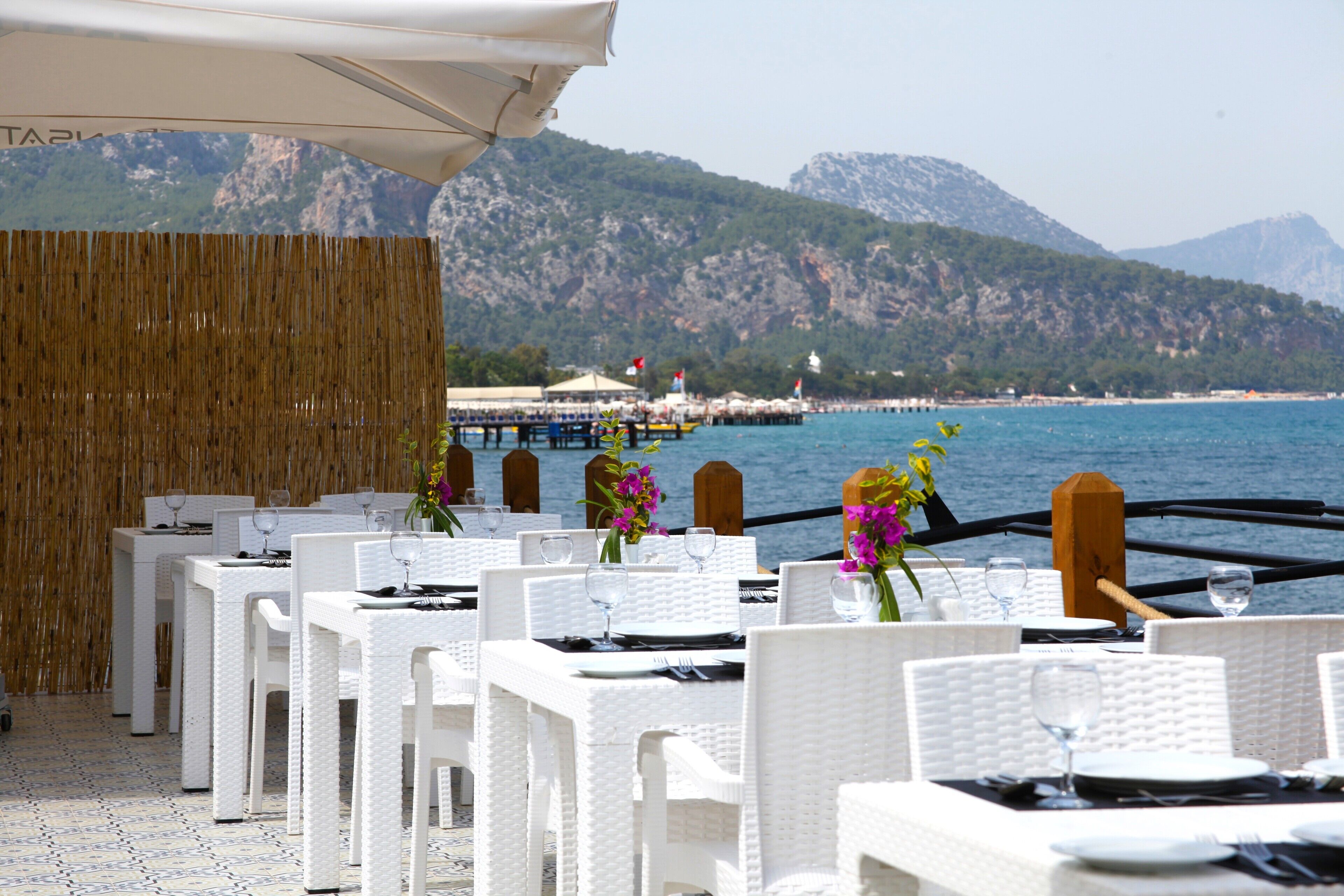 A seaside restaurant with white wicker tables and chairs, set against a backdrop of mountains and the sea.