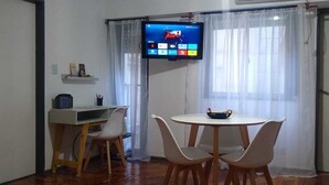 Ein kleines Zimmer mit einem an der Wand befestigten Fernseher, einem Schreibtisch mit Stuhl, einem runden Tisch ebenfalls mit Stuhl und einem Fenster mit transparenten Vorhängen.