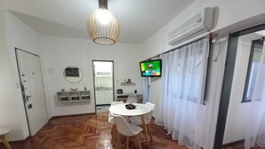 Ein Zimmer mit Holzboden im Fischgrätmuster, einem runden weißen Tisch mit Stühlen, einem an der Wand montierten Fernseher und einer zur Küche führenden Tür.