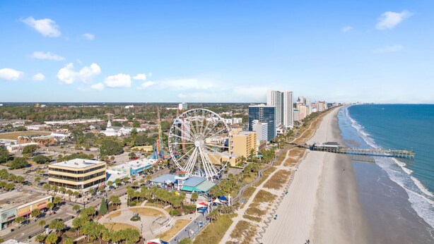 Vista aerea di una città costiera con una ruota panoramica ben visibile, spiaggia e un molo che si protende sull'oceano.