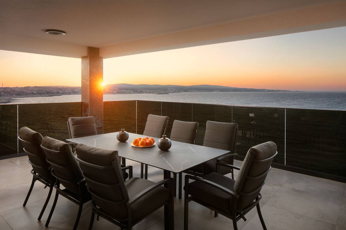 Ein Essbereich mit Tisch und Stühlen, einer Obstschale und Blick auf den Sonnenuntergang über dem Wasser.