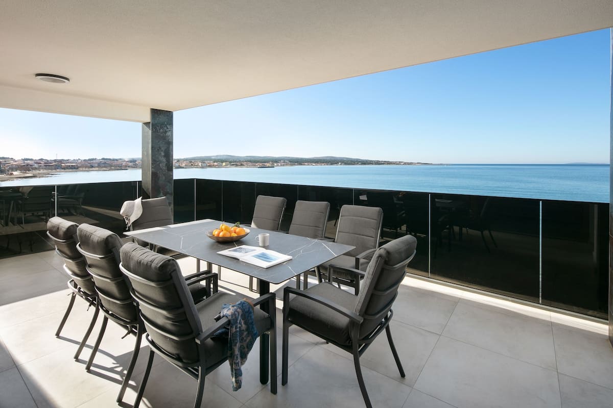 Ein moderner Essbereich im Freien mit Panoramablick auf das Meer, einer Glasbrüstung und zeitgemäßer Möbel.