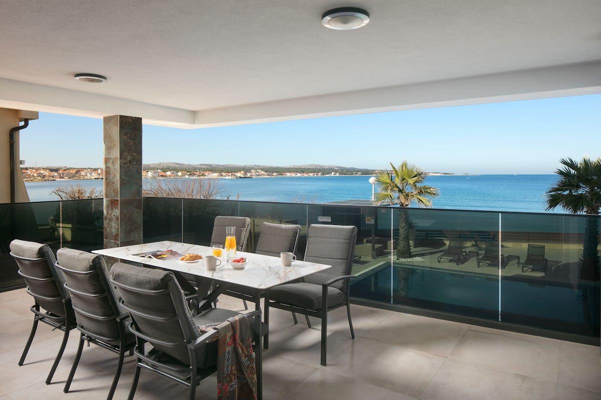 Ein Balkon mit Tisch und Stühlen, der Blick auf ein Gewässer und eine entfernte Küstenlinie freigibt.
