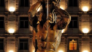 Eine bronzene Statue einer muskulösen Figur in wallendem Gewand, vor einer Gebäudeansicht mit beleuchteten Fenstern.