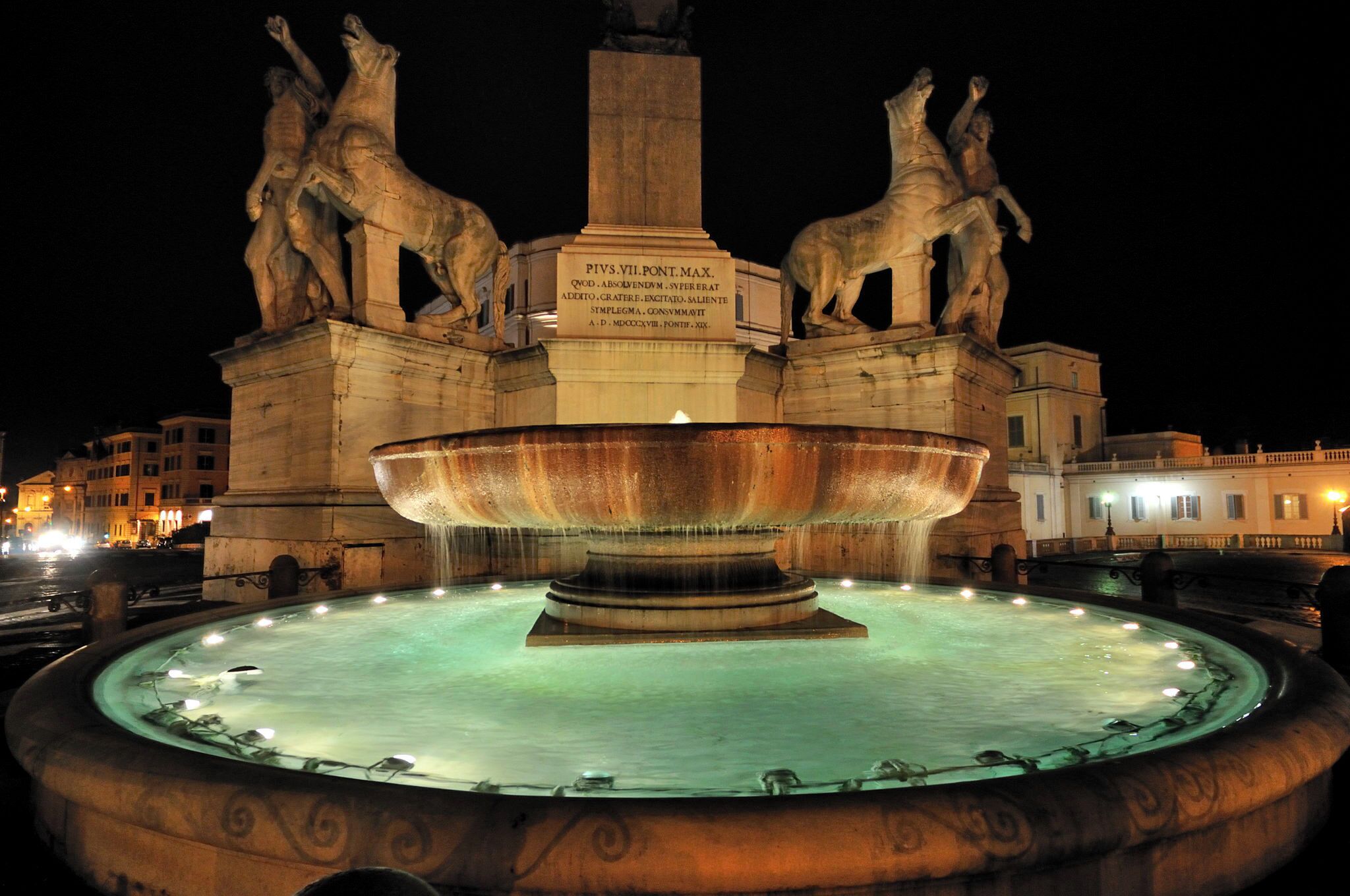 A nighttime view of a historic fountain with statues and a central basin, surrounded by buildings.