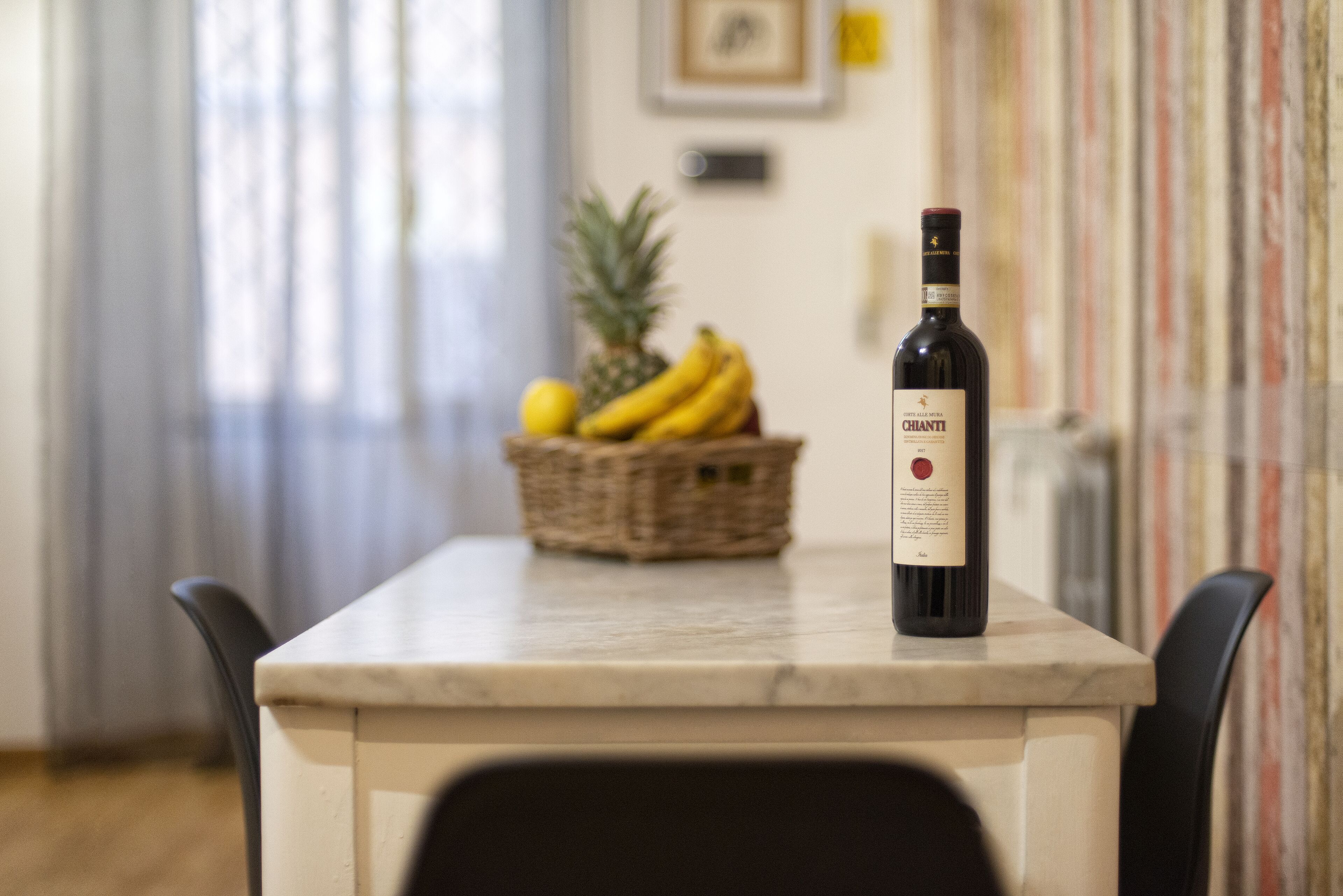 Una botella de vino Chianti sobre una mesa de mármol, con una cesta de mimbre que contiene plátanos y una piña al fondo.