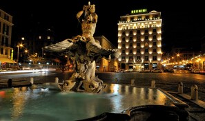 Vista nocturna de una fuente con una estatua de león en el centro, rodeada por una plaza adoquinada con edificios al fondo, incluido un hotel con 'BERNINI' iluminado en su fachada.