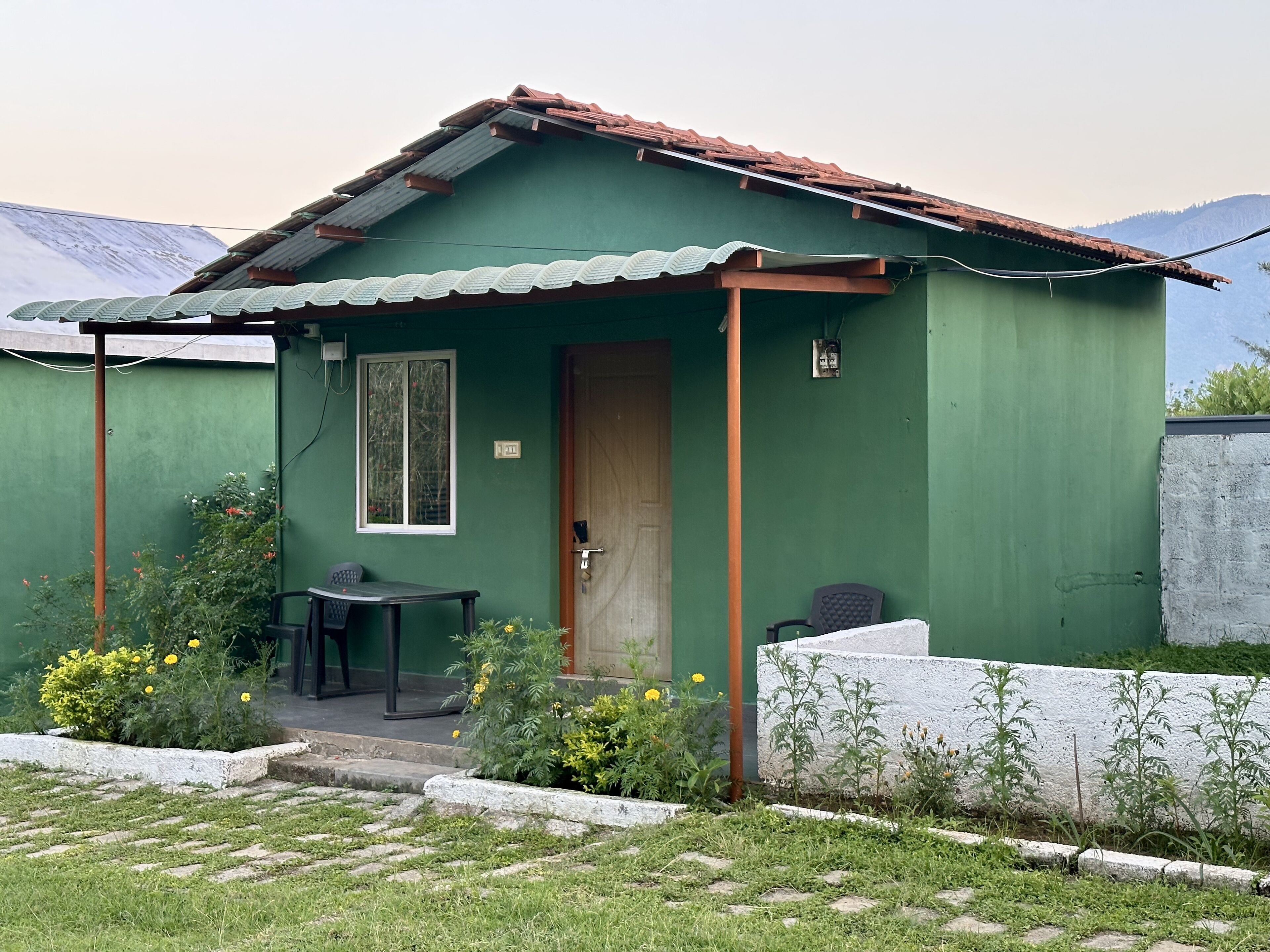 A small, single-story building with a green exterior and a corrugated metal roof. There is a door with a window, a small table, and a chair outside. The building is surrounded by a low stone wall and has a grassy area with yellow flowers in front.