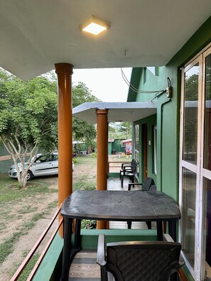 Ein überdachter Außenbereich mit Tisch und Stühlen, Blick auf ein geparktes Auto sowie ein grünes Gebäude mit einem Fenster.