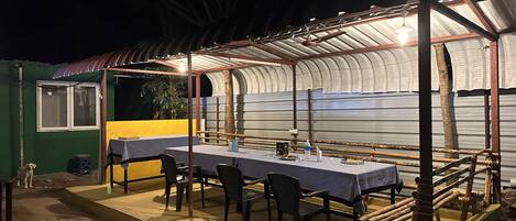 Un espace extérieur aménagé pour prendre les repas, avec une table et des chaises sous un auvent, une petite table avec une chaise, et un chien à proximité.